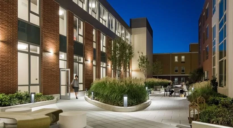 Nighttime scene of an outdoor courtyard between modern buildings, with seating areas, plants, and people sitting and walking.