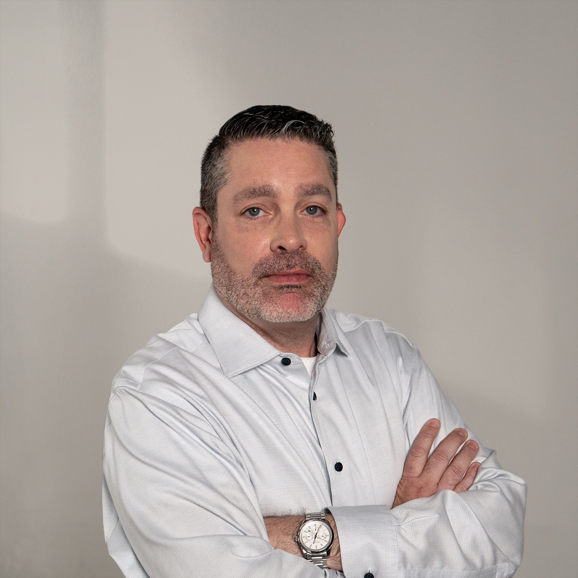 Man with dark hair and beard wearing a white dress shirt, standing with arms crossed against a plain background.