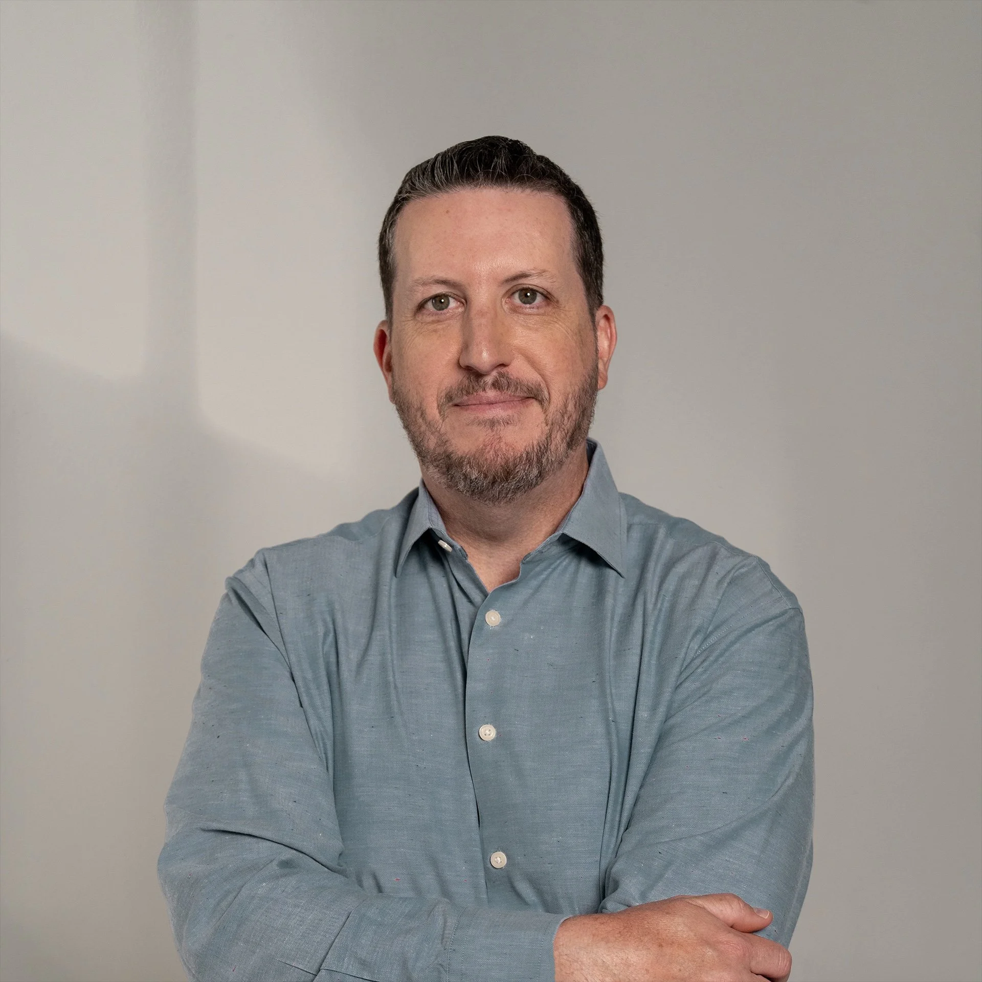 A man with dark hair and a beard, wearing a gray button-up shirt, looking at the camera with a neutral expression, standing against a plain background.