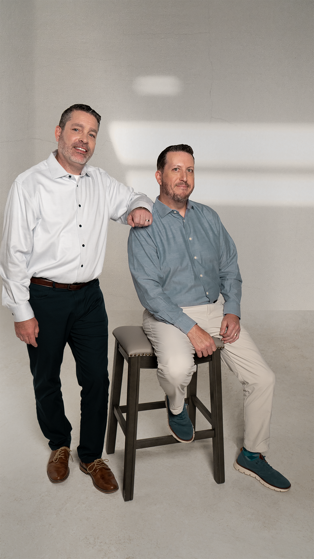 Two men in professional attire, one standing with his arm resting on the shoulder of the other who is sitting on a stool, in a neutral setting with plain background.