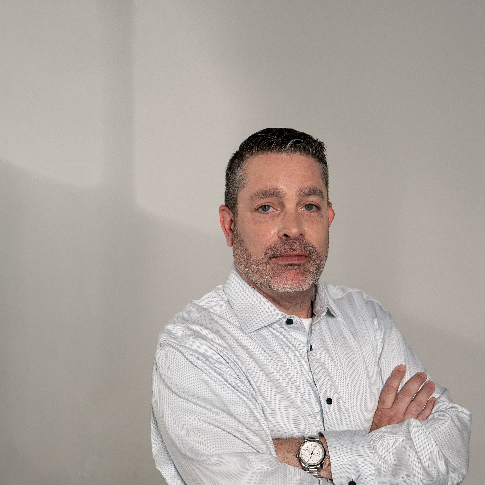 A man with short, dark, slicked-back hair and a gray beard, wearing a light gray dress shirt and a silver watch, standing against a plain, light-colored wall. He is crossing his arms and looking directly at the camera.
