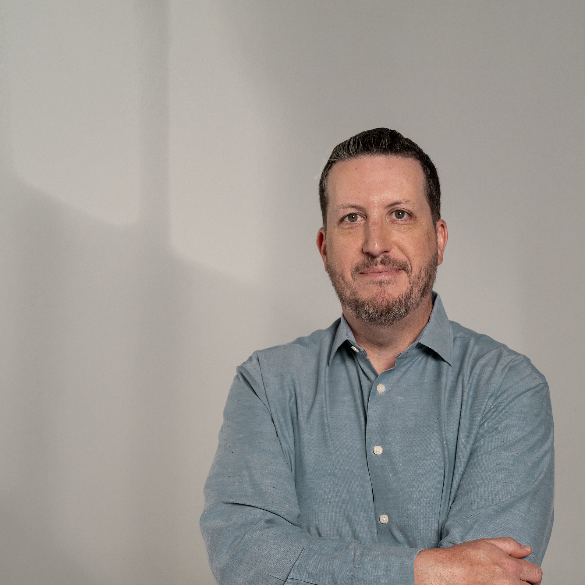 A man with short dark hair, a beard, and gray eyes, wearing a light gray button-up shirt, standing against a plain background.