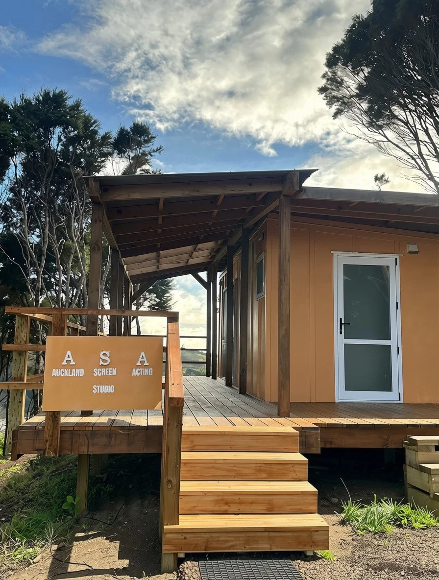 Wooden deck with stairs leading to a small building with glass door, positioned outdoors among trees under a partly cloudy sky, with a sign reading "A S A Auckland Screen Acting Studio."