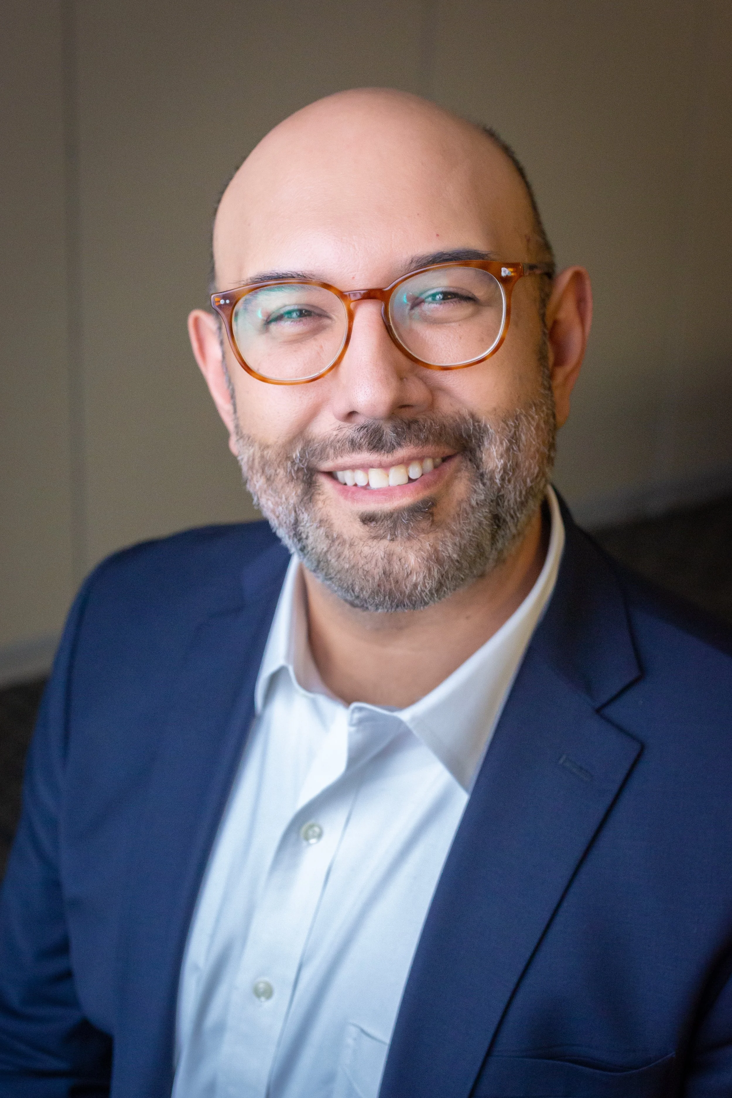 Close-up of Ruben Gonzales with glasses, bald head, beard, wearing a navy suit and white shirt, posing indoors.