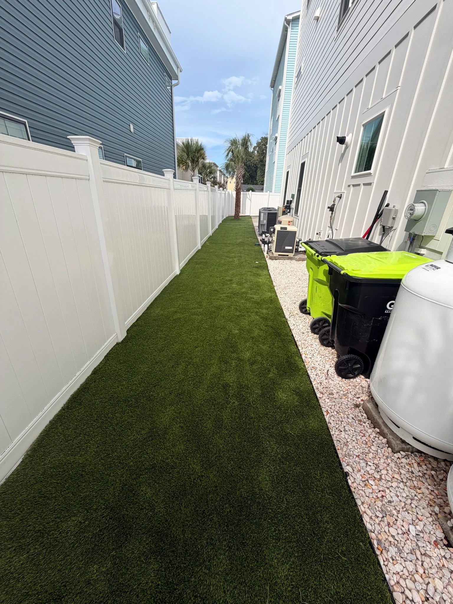 Narrow side yard with artificial turf, white privacy fence, palm trees, HVAC units, and outdoor trash bins between two homes