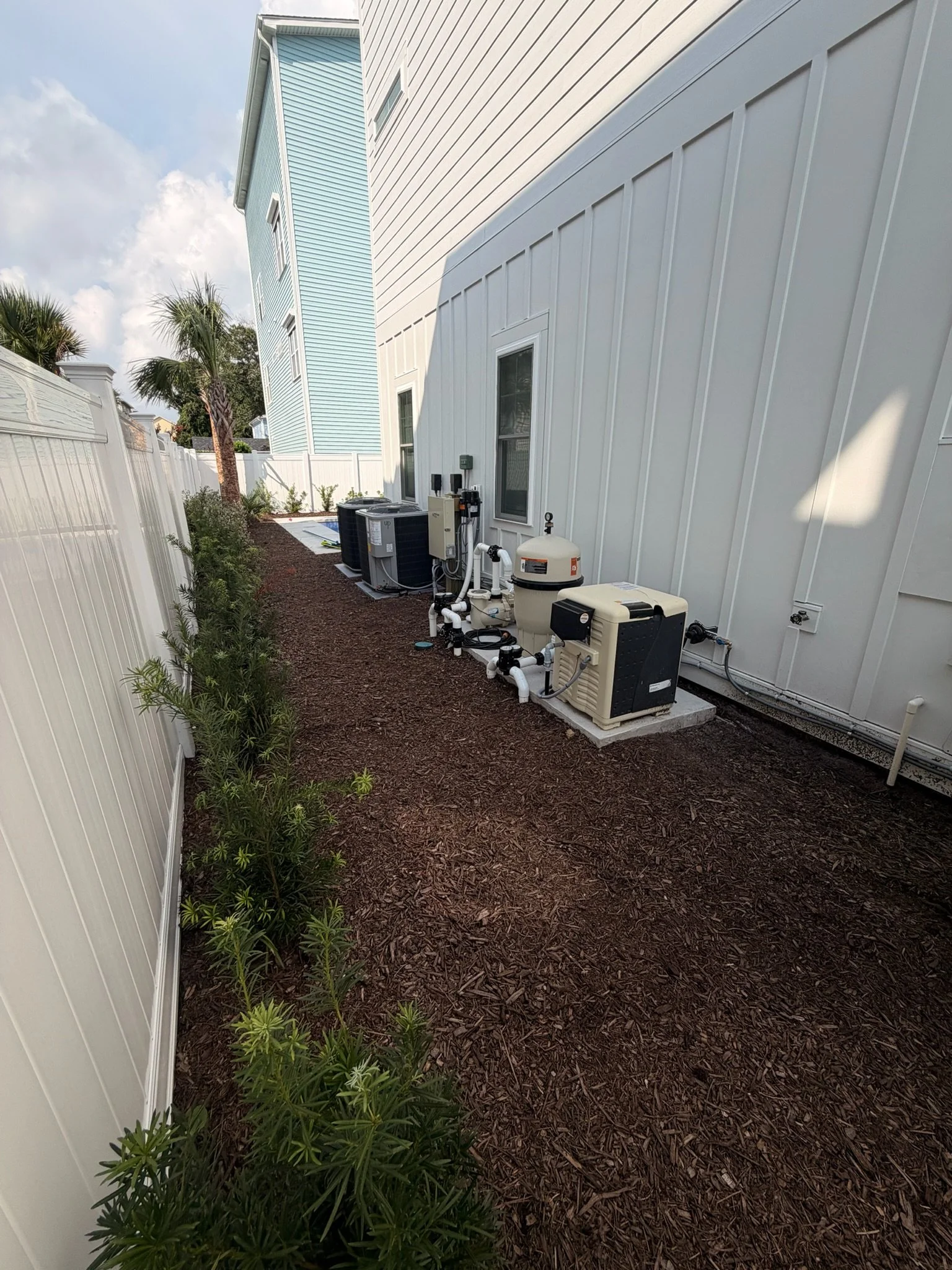 Side yard with mulch landscaping, white privacy fencing, palm trees, and outdoor pool equipment beside a modern home