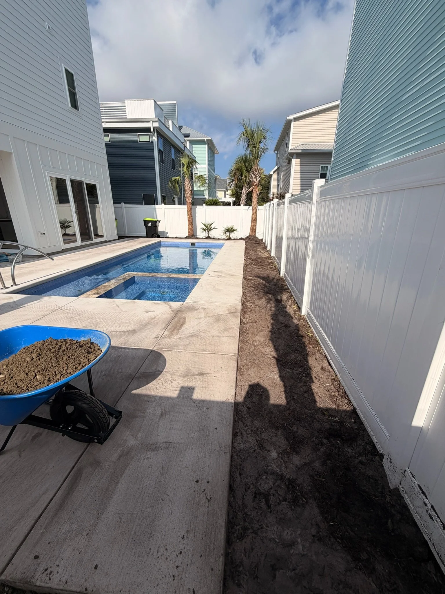 Backyard with a rectangular swimming pool, palm trees, white fencing, modern homes, and a wheelbarrow filled with soil.