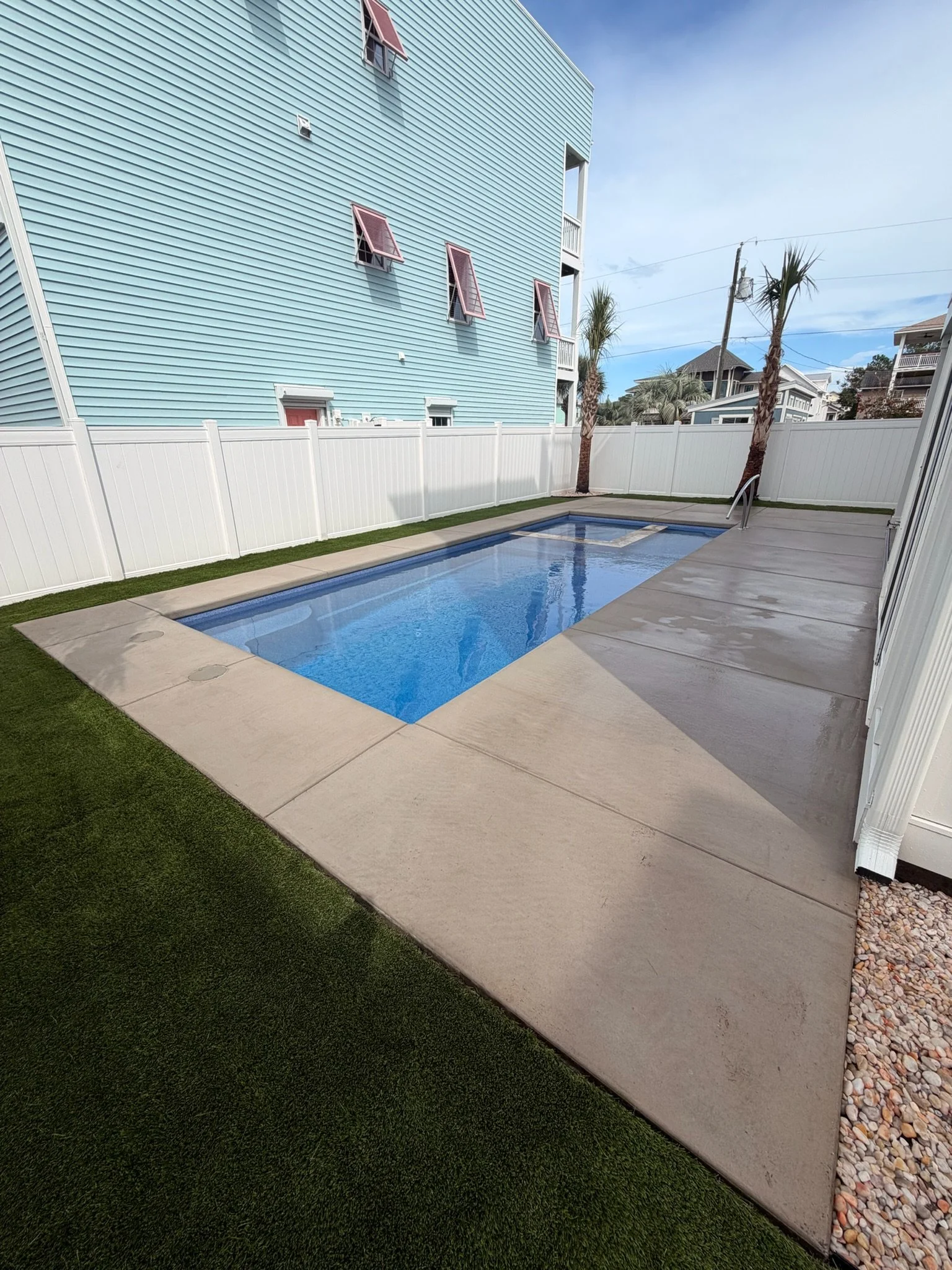 Backyard with a rectangular swimming pool, concrete patio, artificial turf, palm trees, and white privacy fencing beside a beach-style home.
