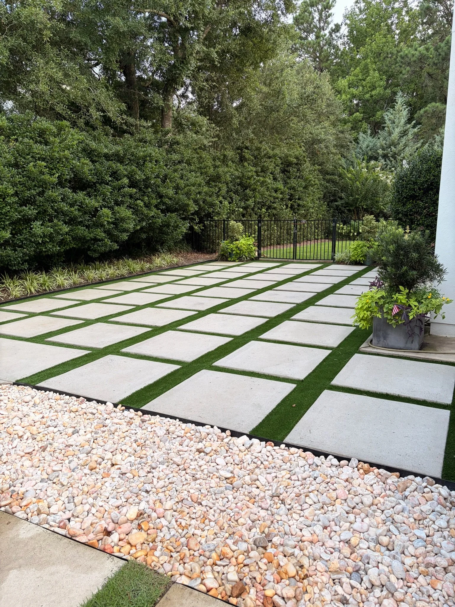 Landscaped backyard with large concrete stepping stones, artificial grass, decorative river rocks, lush shrubs, potted plants, and a black metal gate