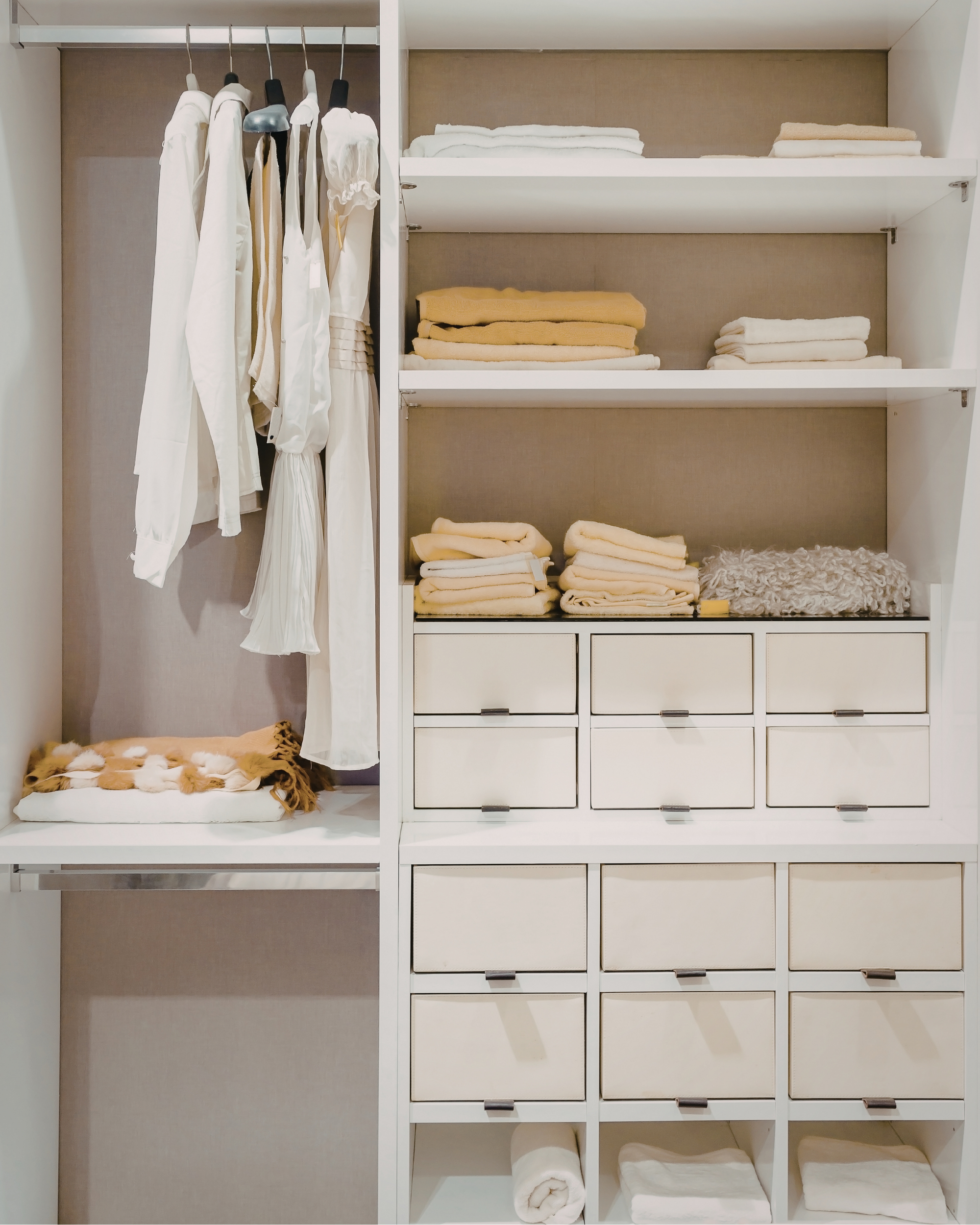 White closet with hangers holding white and cream clothing on the left and shelves on the right filled with folded towels, blankets, and a rolled-up towel at the bottom right.
