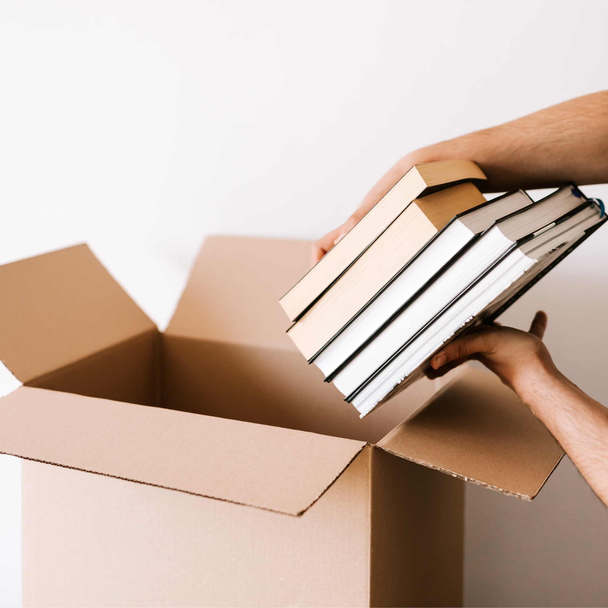 A person's hand holding a stack of books inside a cardboard box on a beige surface.