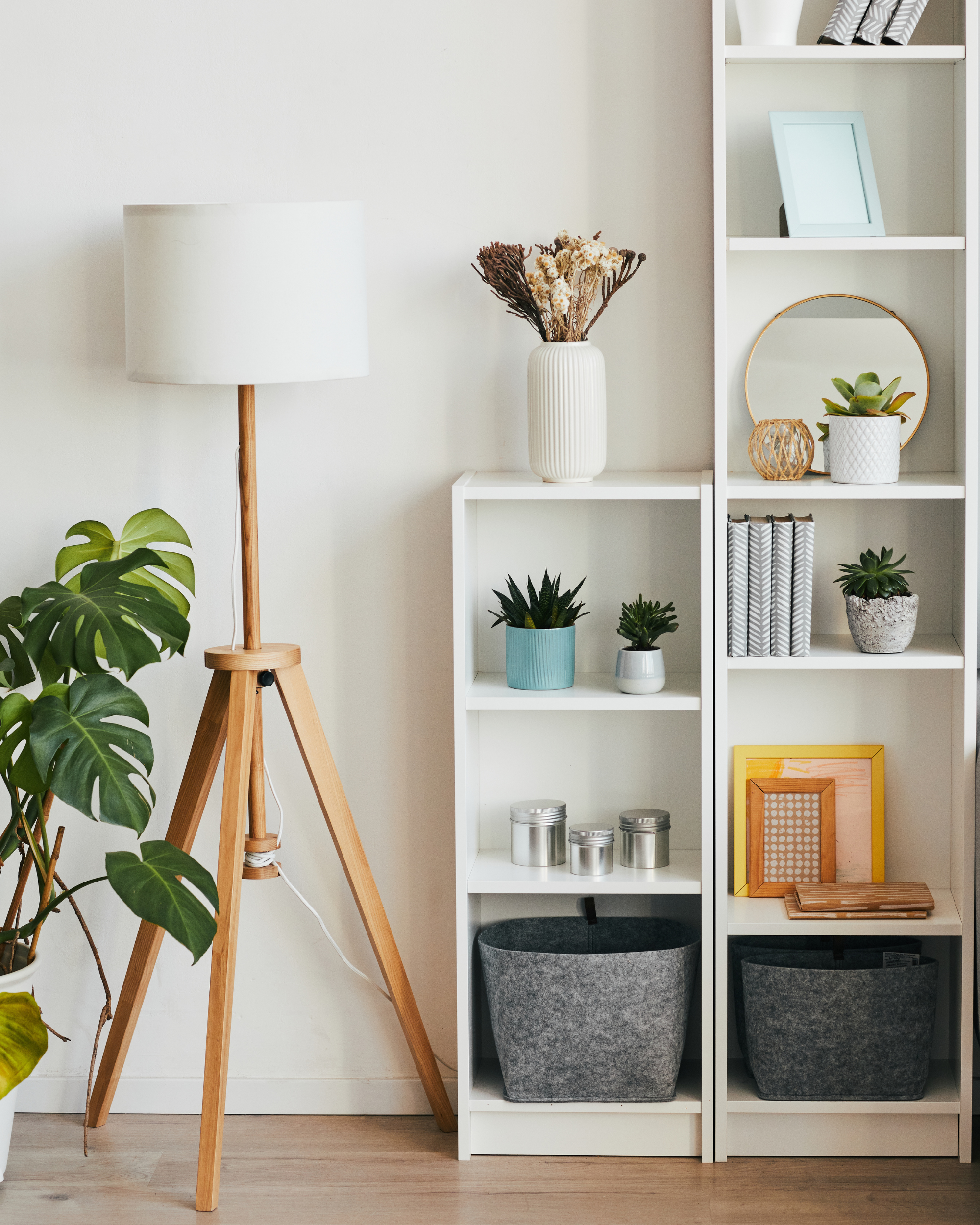 Interior view of a room with a white bookshelf filled with decorative objects and plants, a grey fabric storage bin, a wooden floor, and a wooden tripod lamp with a white shade. A large leafy green plant is visible in the lower left corner.