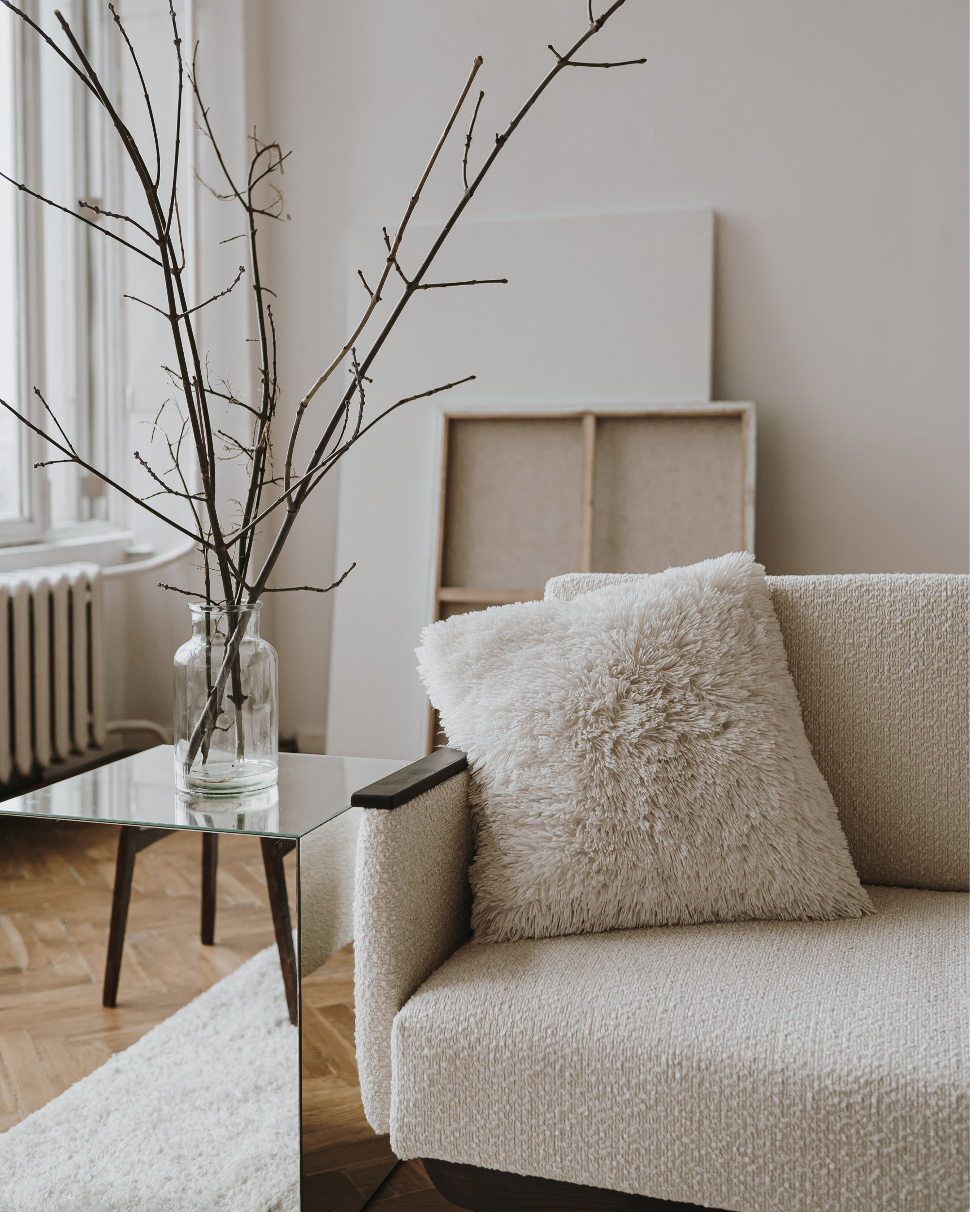 A living room corner with a beige upholstered sofa, a fluffy white pillow, a glass side table with pine branches in a clear vase, a white rug, and a blank white wall with a partially opened empty picture frame.