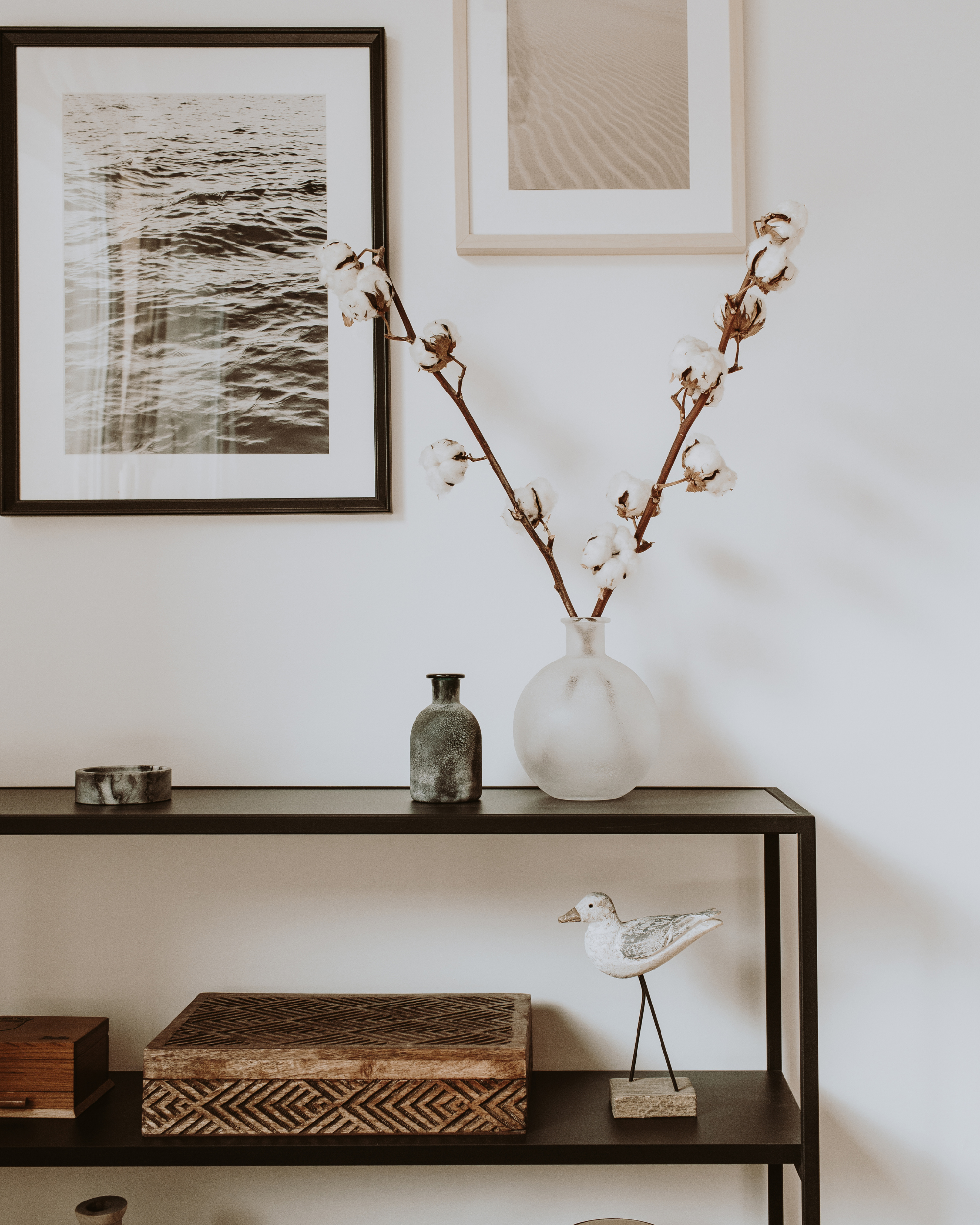 A black metal shelf with decorative items and framed artwork on white walls. The top shelf has two framed black-and-white photographs of water and sand. Centered is a large white vase with cotton stems. On the lower shelf, there are two small vases, a geometric wooden tray, and a white ceramic Boston duck figurine. The middle shelf includes a small gray vase and a patterned wooden box. The wall features a photo of water and another of sand.