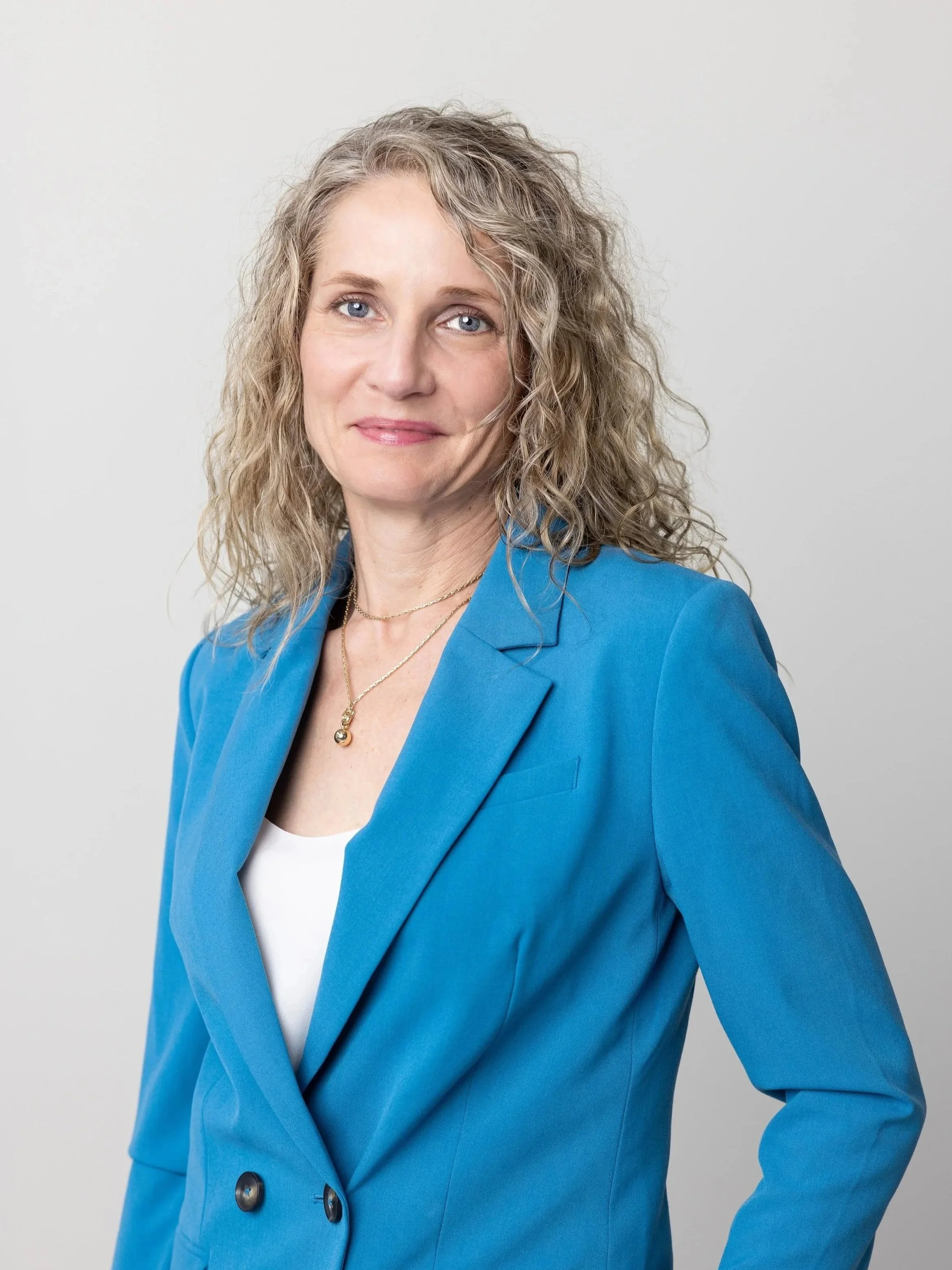 A woman with curly blonde hair wearing a blue blazer over a white top and gold jewelry, standing against a light gray background.