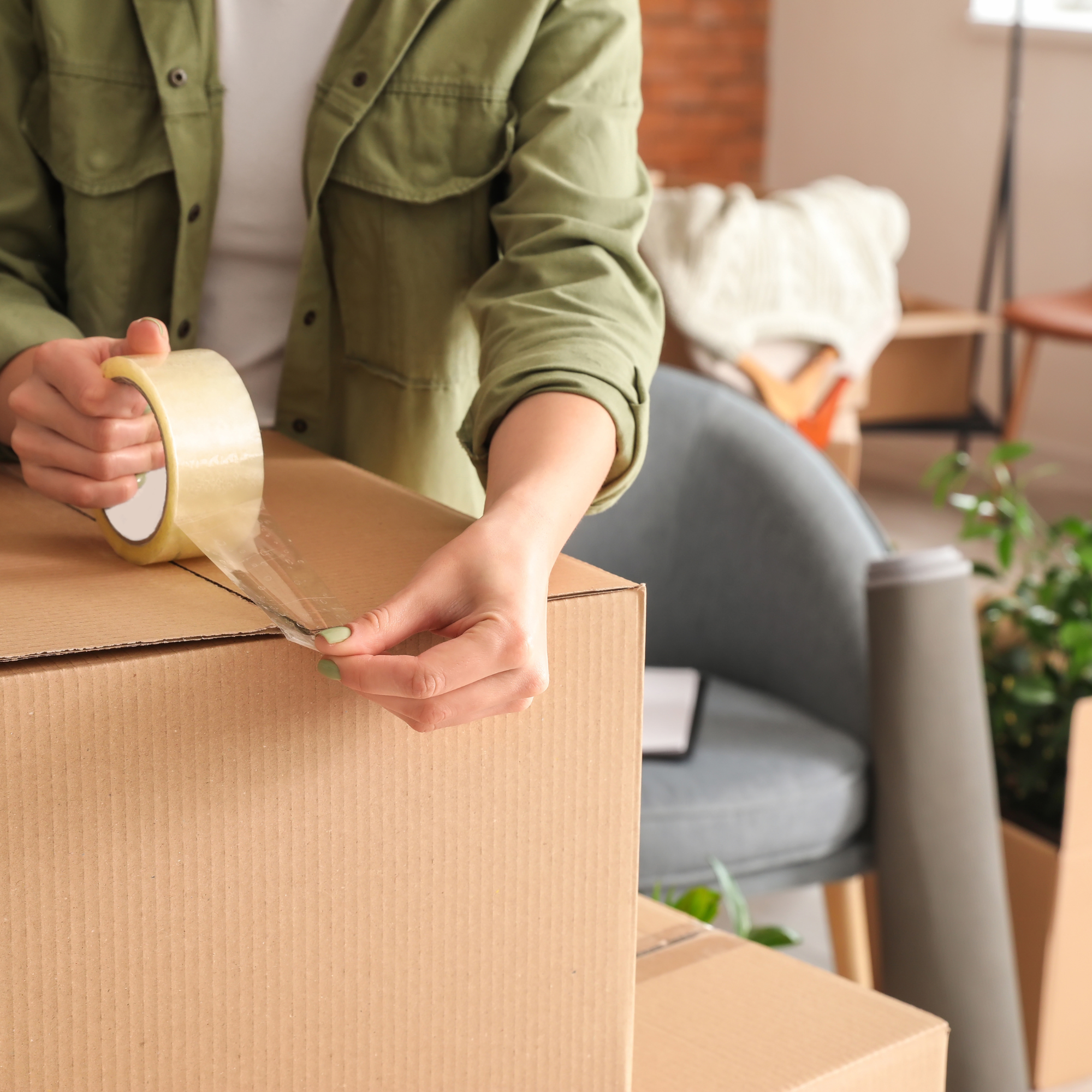 Close-up of a person's arm and hand holding a roll of adhesive tape, partially unrolled, over a cardboard box on a wooden table in a room with indoor plants and furniture.