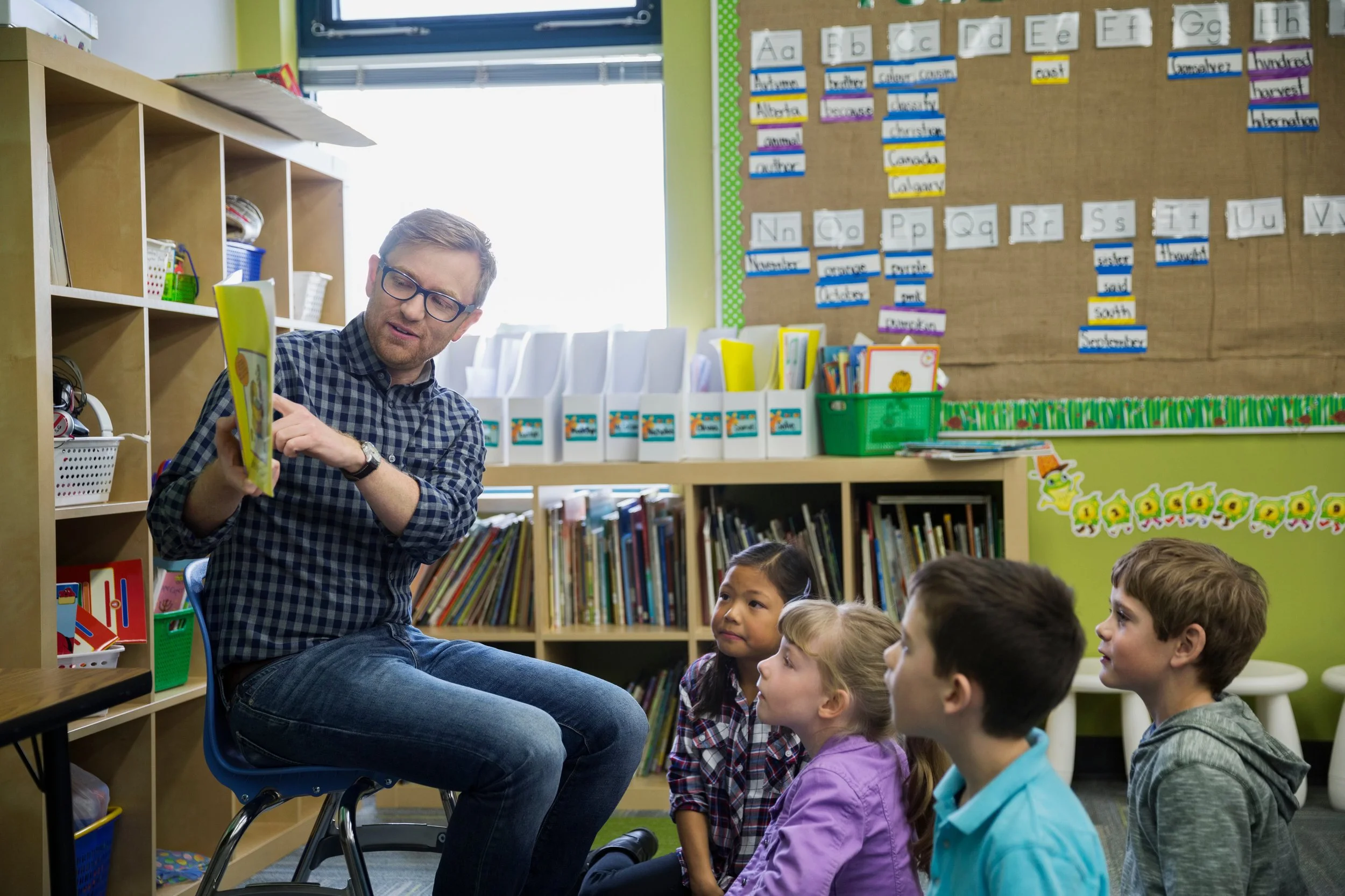 A teacher reads a book to a small group of elementary school children seated on the floor in a classroom library.