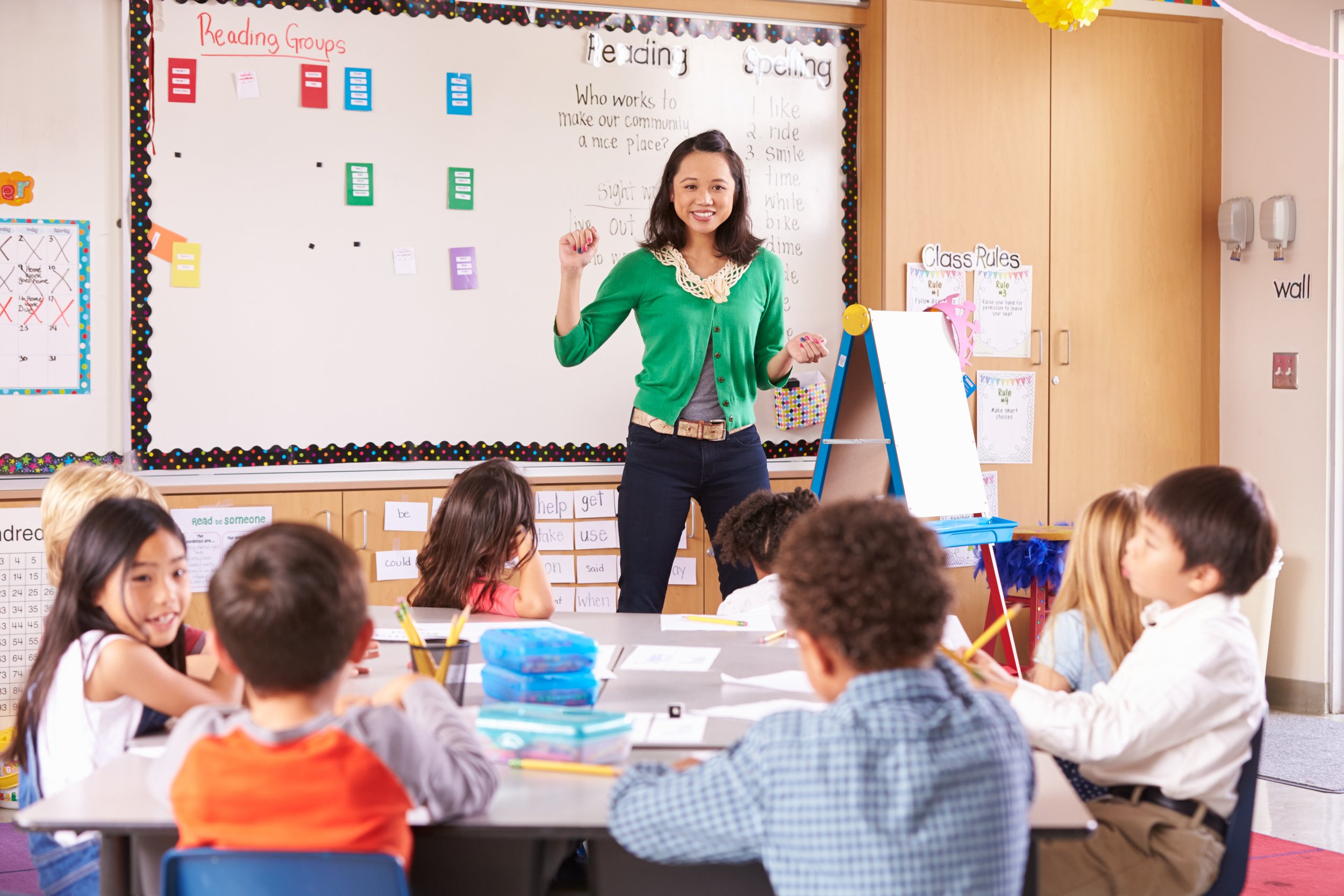 A teacher leads a lesson at the front of an elementary classroom while students sit at tables and participate.