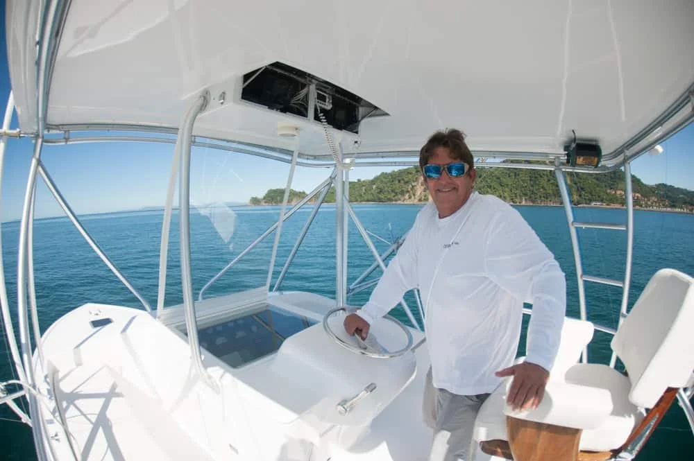 Man in white shirt and sunglasses steering a boat on the water with land in the background