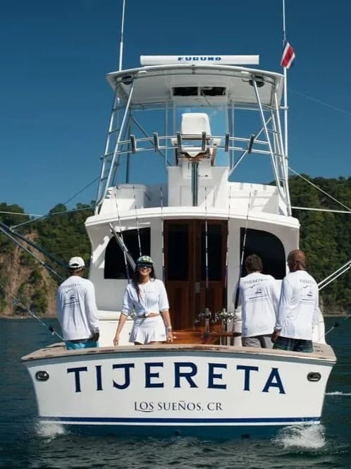 Four people are on a boat named Tijereta, with a woman smiling at the camera and three others with their backs turned. The boat is on water with a green hillside in the background.