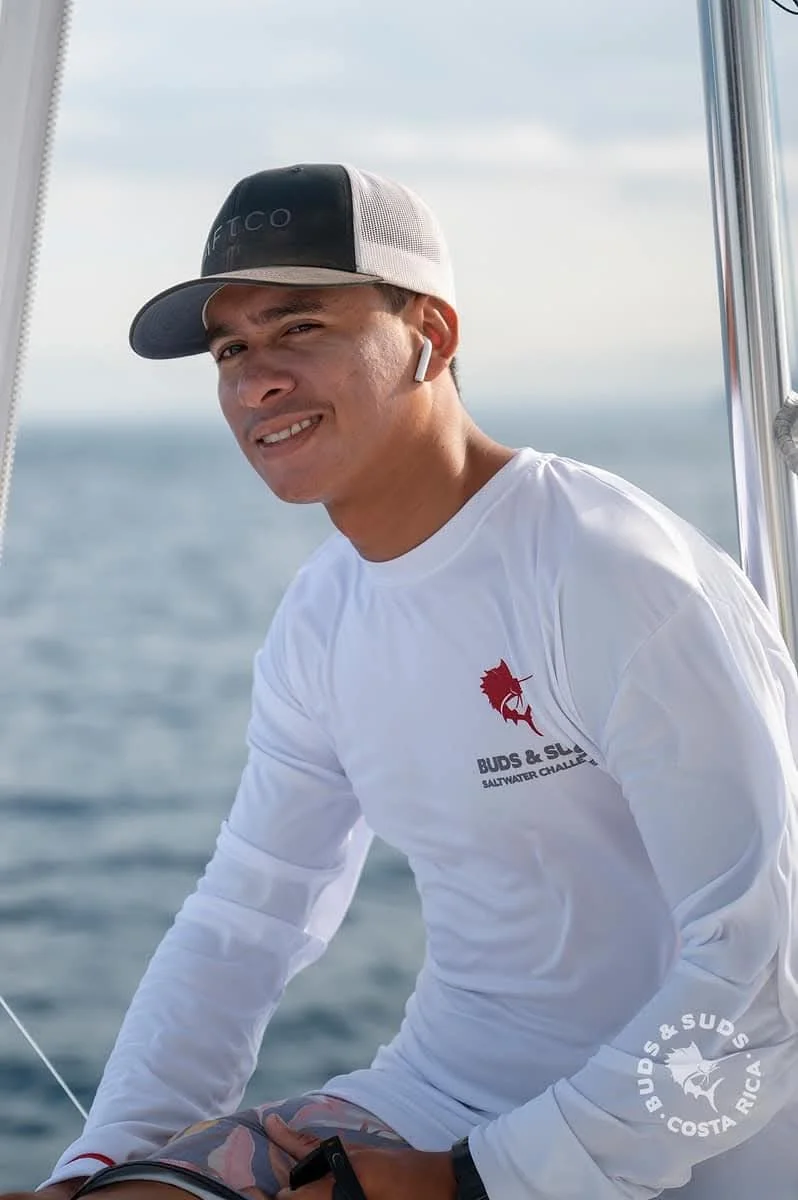 A person in a white shirt and wide-brimmed hat fishing on a boat in the ocean. A dolphin is jumping out of the water in the distance.