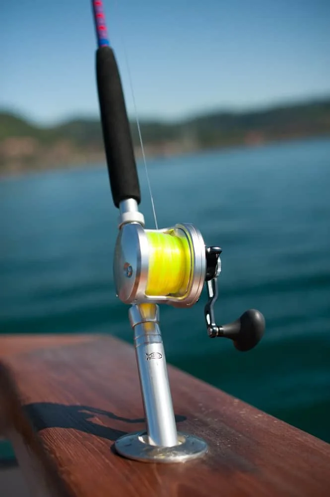 A fishing rod with yellow line mounted on a wooden boat railing near a body of water, with a blurred landscape in the background.