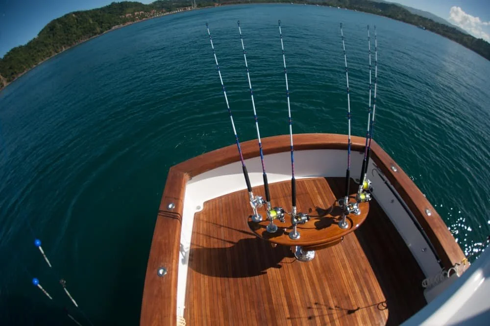 A boat with fishing rods on the deck, out on a body of water near a hilly shoreline.