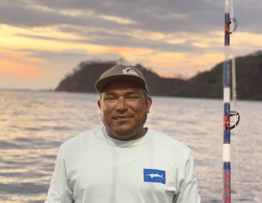 Man standing on a boat at sunset, smiling, with a fishing rod nearby and a landmass in the background.
