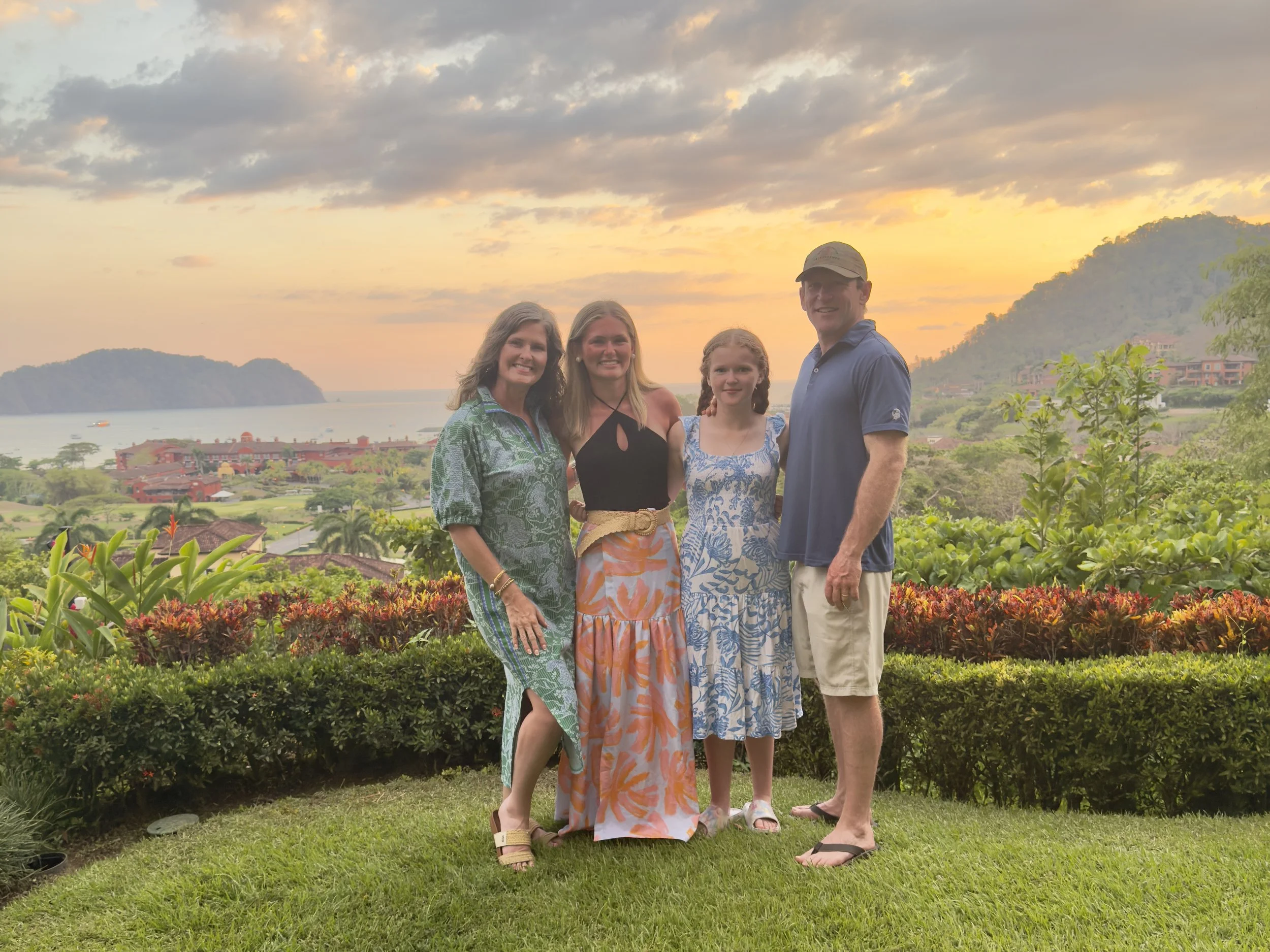A family of four standing outdoors on a grassy area with a scenic sunset view of water, islands, hills, and buildings in the background