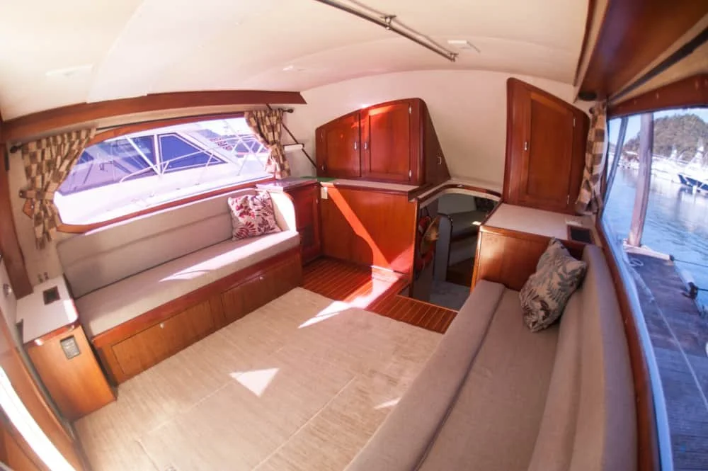 Interior of a boat cabin with wooden furniture, large windows, and seating areas with cushions.