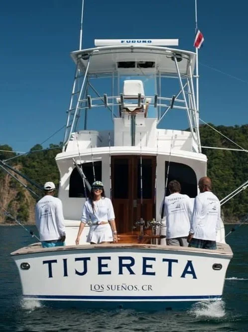 A group of four people on a yacht named Tijereta, docked on calm water with lush green hills in the background. The people are wearing white shirts, and the woman in the front is smiling.