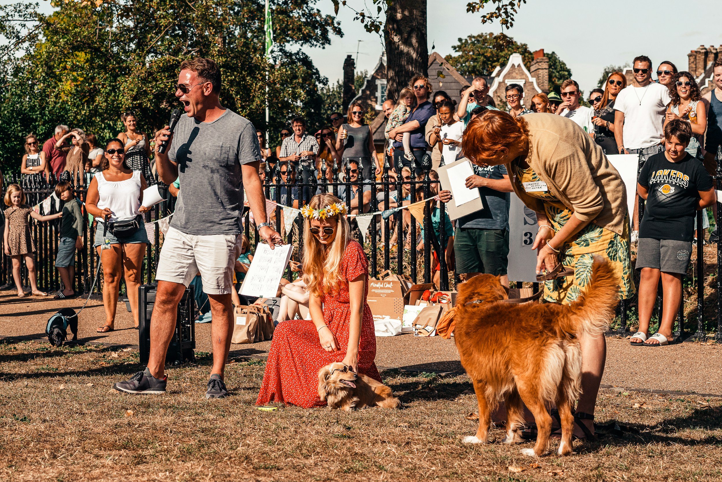A crowd gathered outdoors on a sunny day, watching a man speak into a microphone. A woman with red hair and sunglasses sits on the ground with a small dog, and a larger dog stands nearby. People are smiling, taking photos, and standing behind a black fence, with trees and houses in the background.