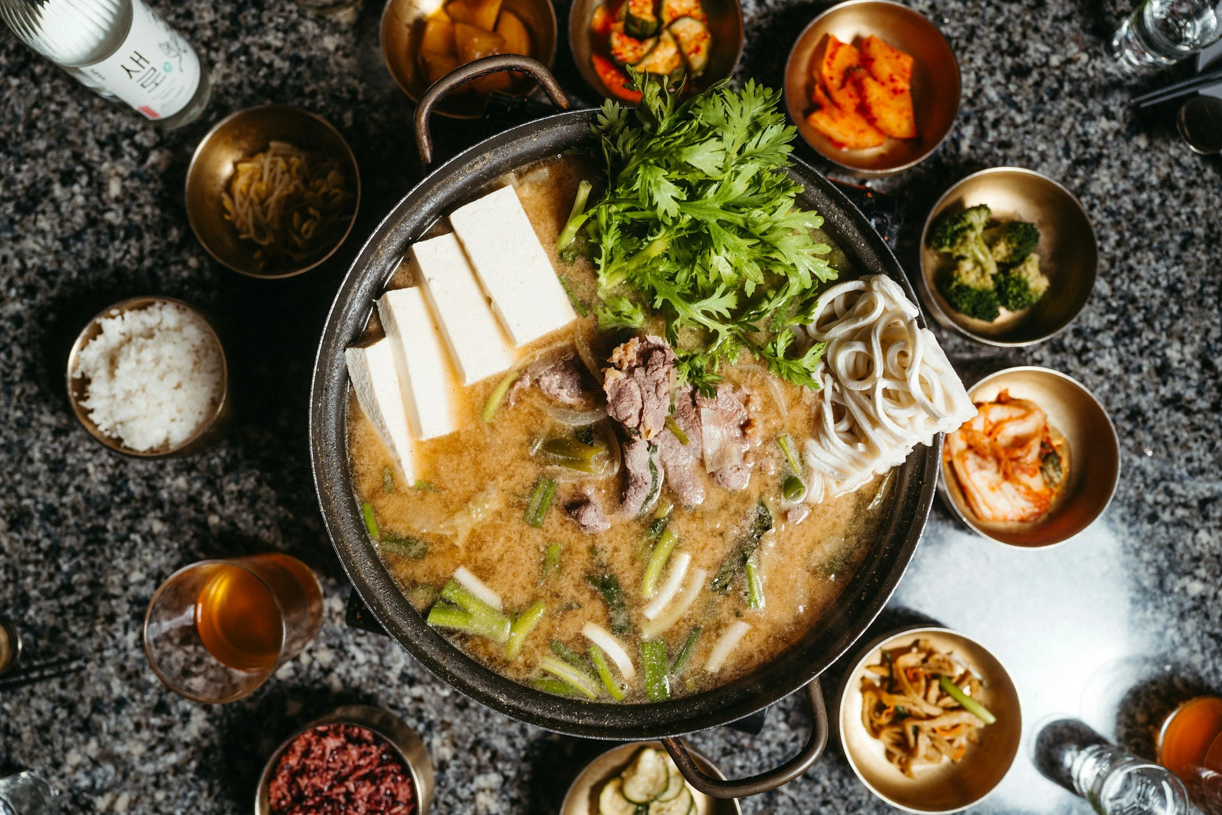 A pot of steaming hot Korean hot pot with tofu, beef, vegetables, and cilantro, surrounded by various side dishes, bowls, and drinks on a granite countertop.