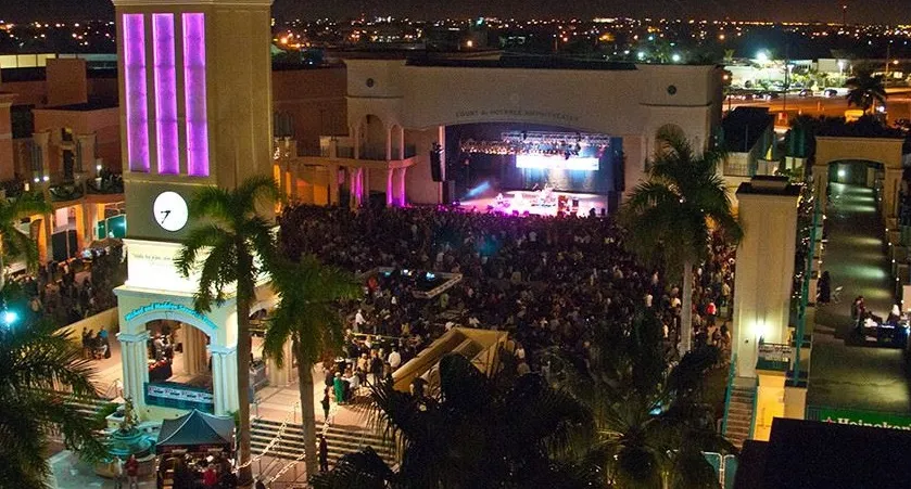 Nighttime outdoor concert in a shopping mall with a stage, large crowd, palm trees, and colorful lighting.