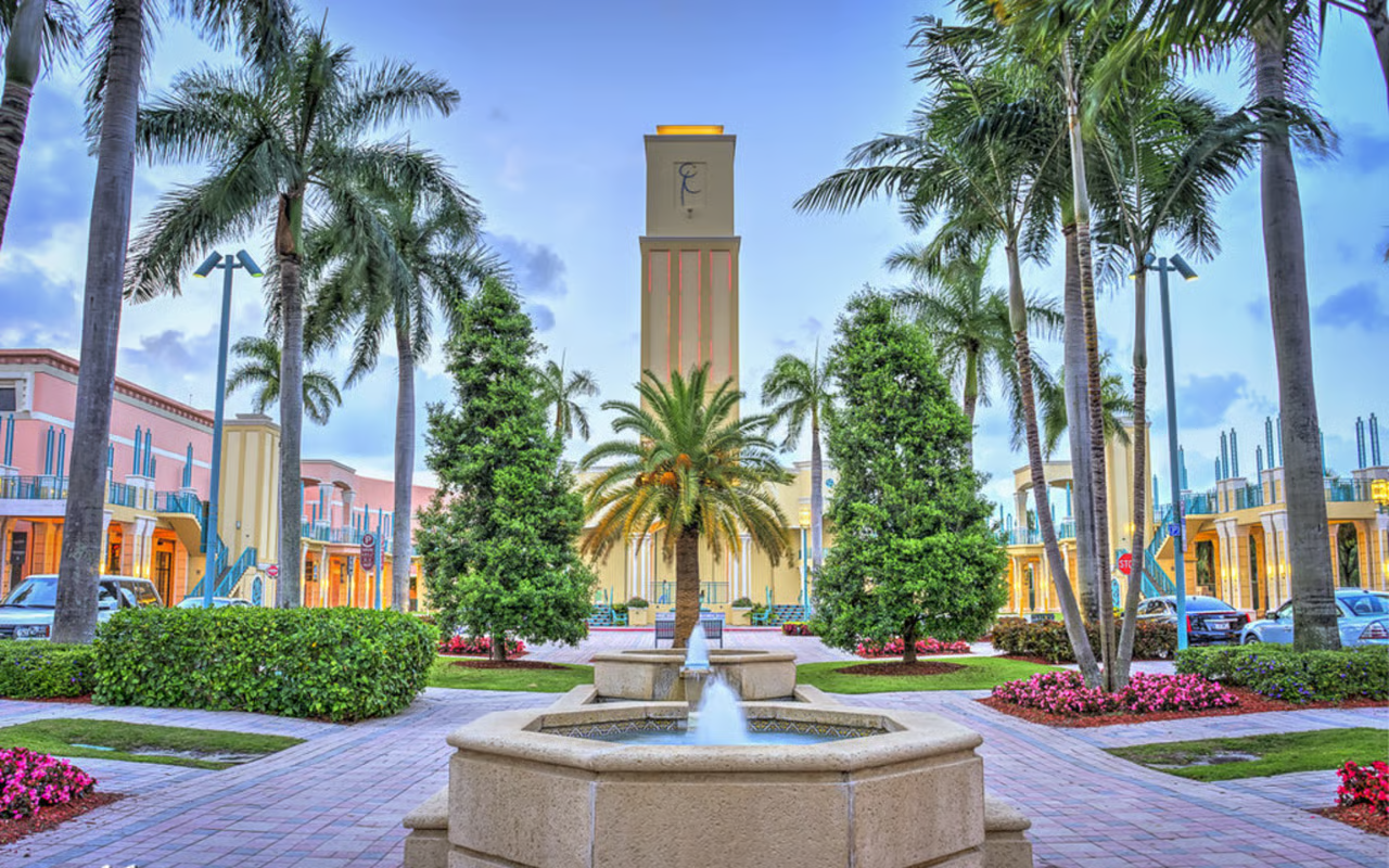 A picturesque shopping center with a central fountain, palm trees, colorful buildings, and parking lot in the background.