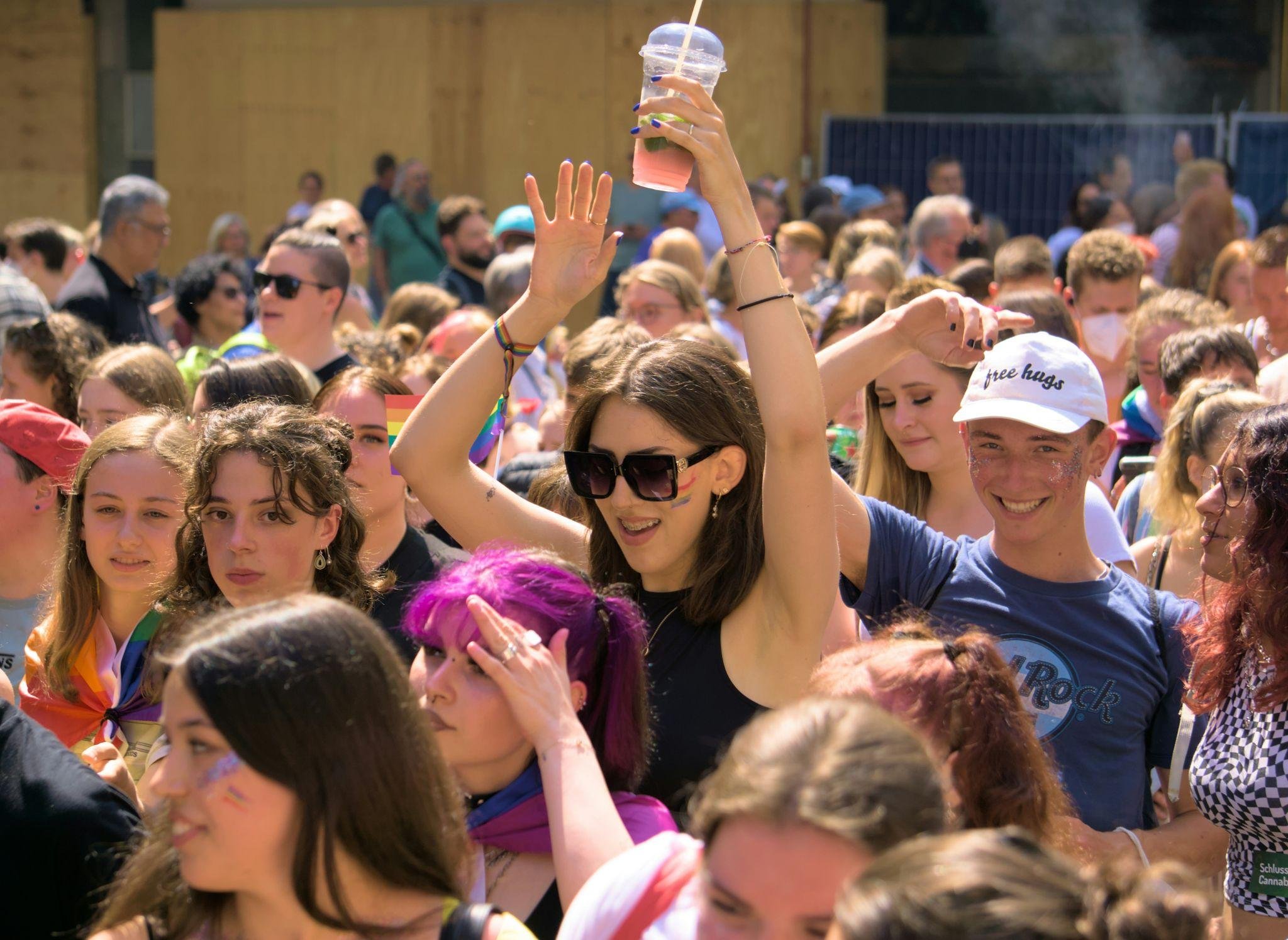 Crowd of people gathered at an outdoor event, some holding drinks. A woman is raising her hand with a drink in it, wearing sunglasses and a rainbow wristband. The scene appears festive with diverse attendees.