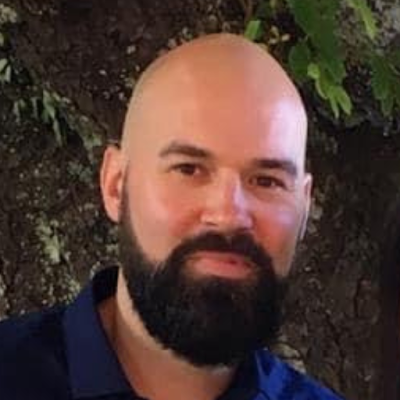 A man with a bald head and a full beard, wearing a navy blue shirt, standing outdoors in front of green foliage.