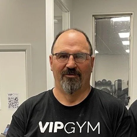 A middle-aged man with glasses and a beard wearing a black VIPGYM T-shirt, standing indoors in front of a door with a window.