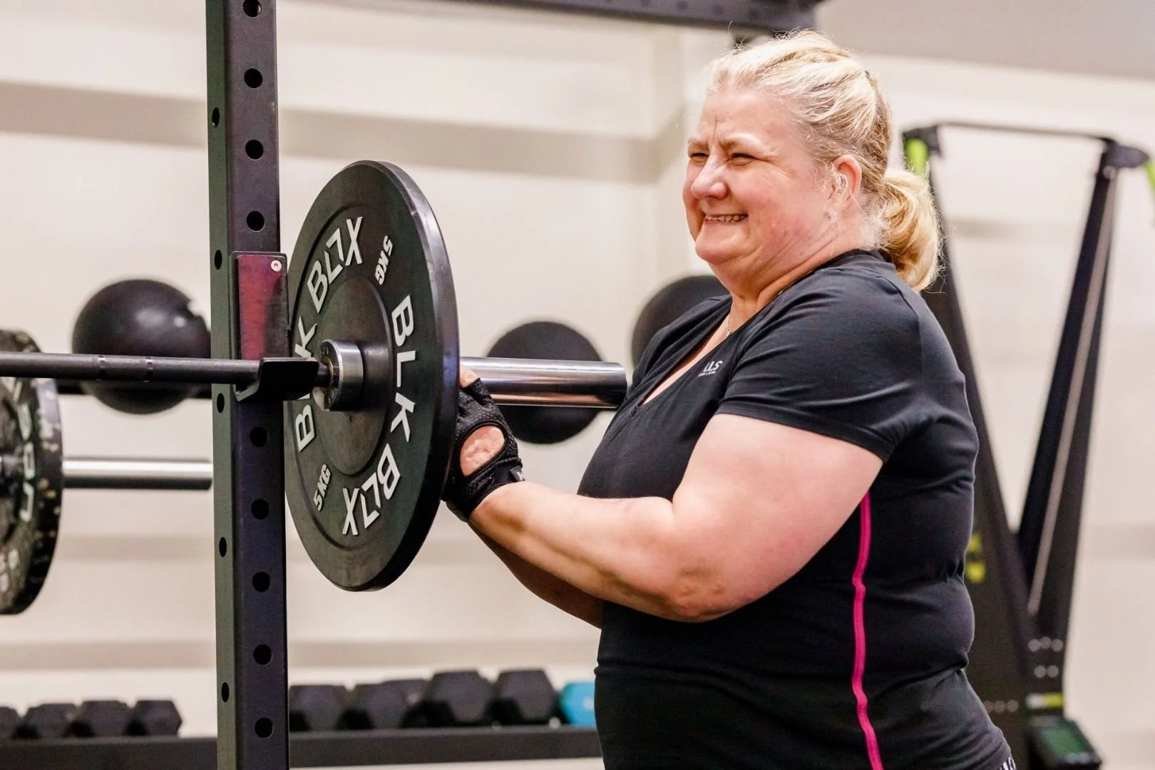A woman lifting a barbell in a gym, smiling with eyes closed, wearing a black workout shirt and gloves, with weight plates on the barbell.