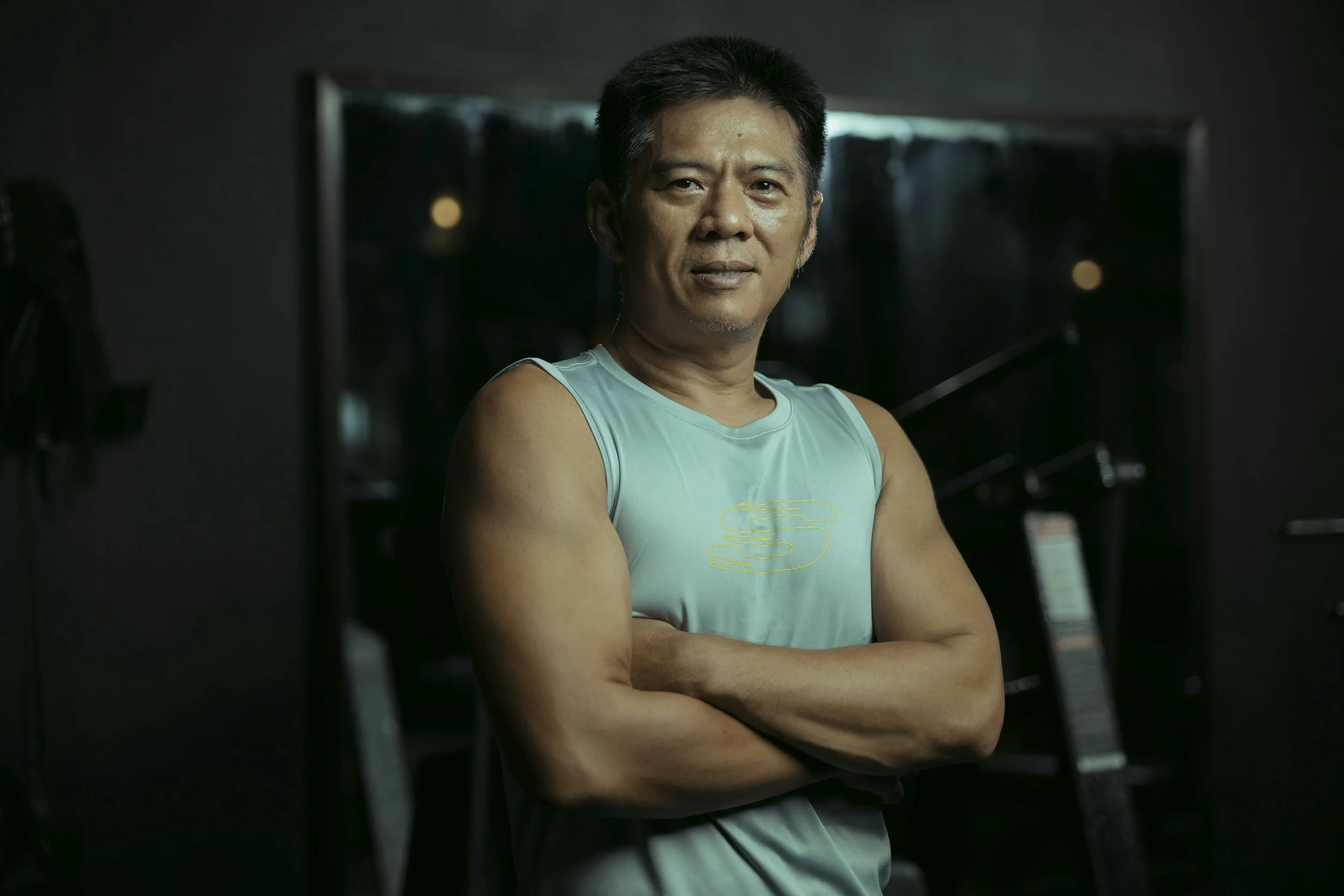 A middle-aged man with short black hair, wearing a light blue sleeveless athletic shirt, stands with his arms crossed in a dark indoor setting. He is looking directly at the camera with a slight smile. The background is out of focus with some faint lights and objects.
