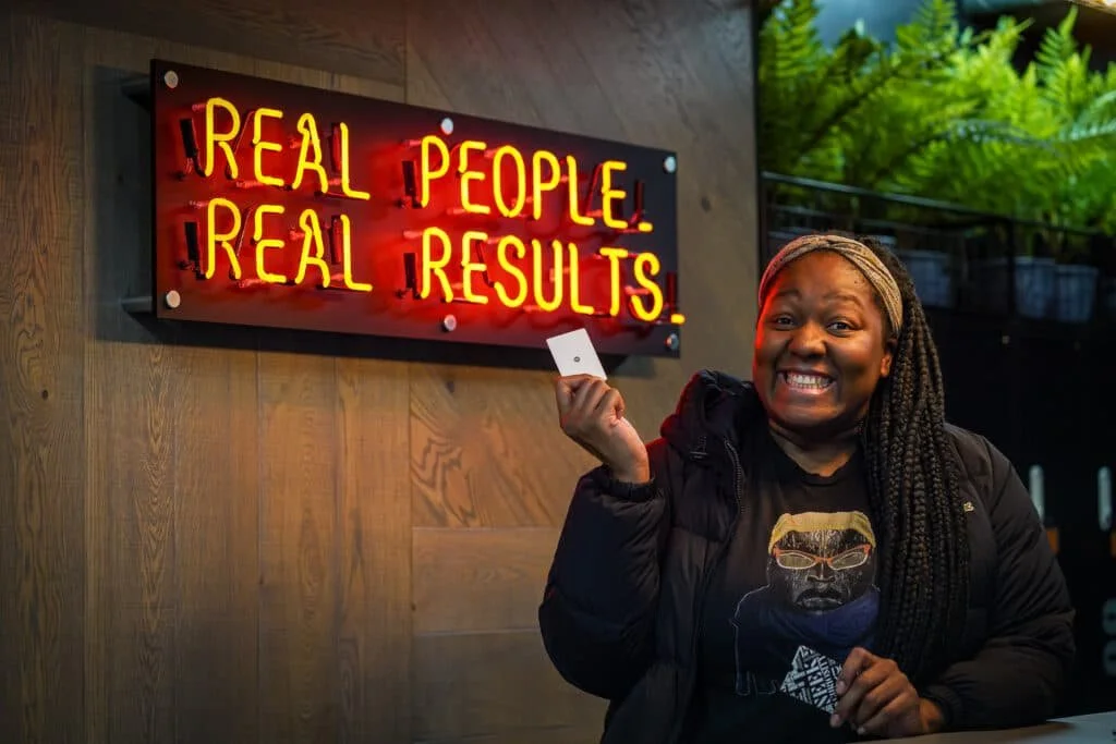 A smiling woman holding a key card in front of a neon sign that says 'Real People Real Results' inside a building.