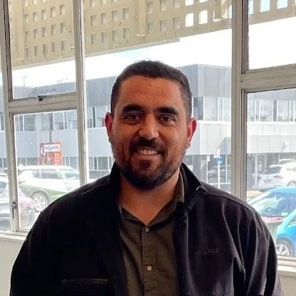 A man with short dark hair and a beard, wearing a dark jacket and a collared shirt, smiling indoors near large windows with cars outside.