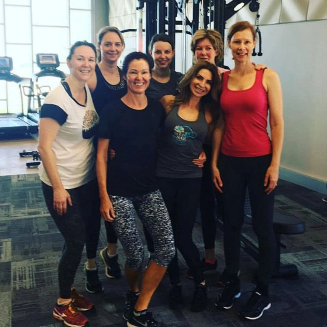 Group of seven women in workout attire posing in a gym with workout equipment in the background.