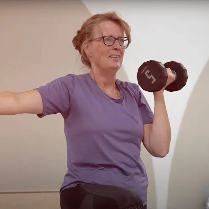 An elderly woman with glasses and red hair, wearing a purple t-shirt, is lifting a 5-pound dumbbell in a room with white walls.