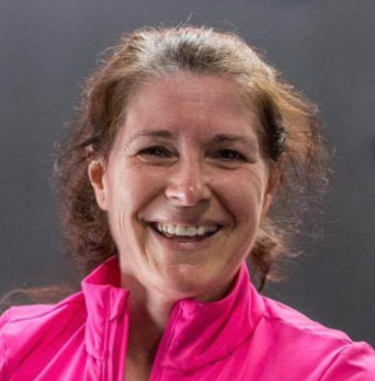 A woman with curly brown hair smiling, wearing a pink jacket, against a dark background.