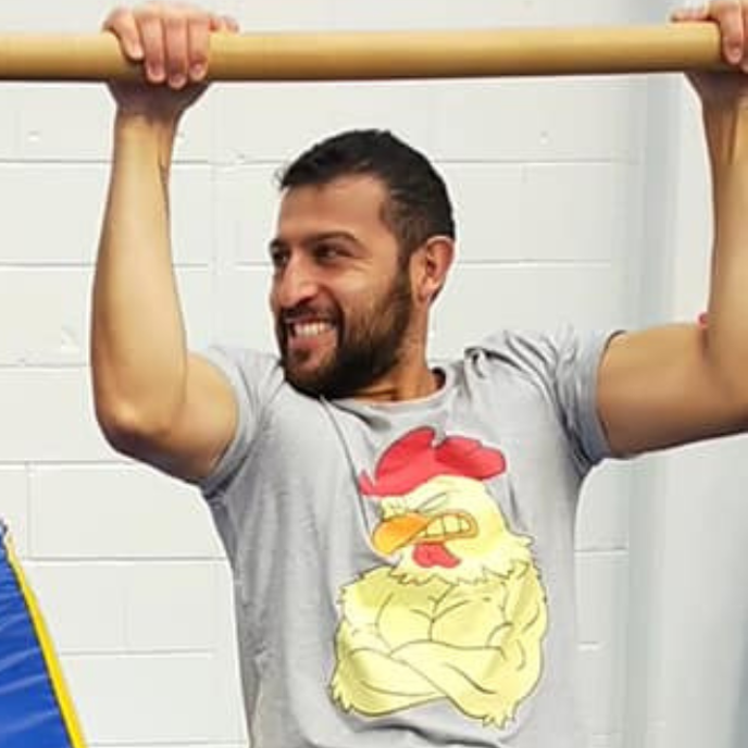 A man hanging from a wooden bar, smiling, wearing a gray T-shirt with a cartoon chicken showing muscles.