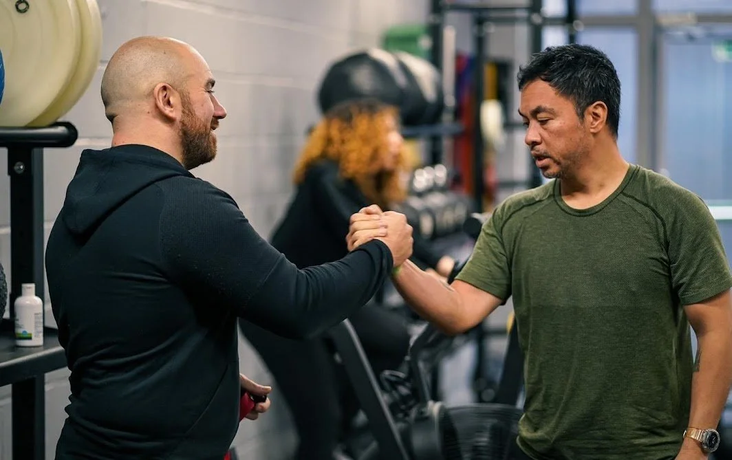 Two men shaking hands in a gym, with gym equipment and a woman exercising in the background.