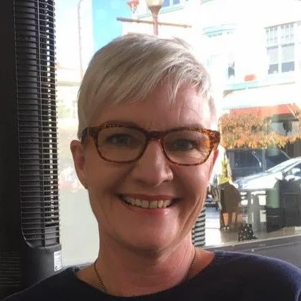 A smiling woman with short blonde hair and glasses, indoors near a window with a city street view.