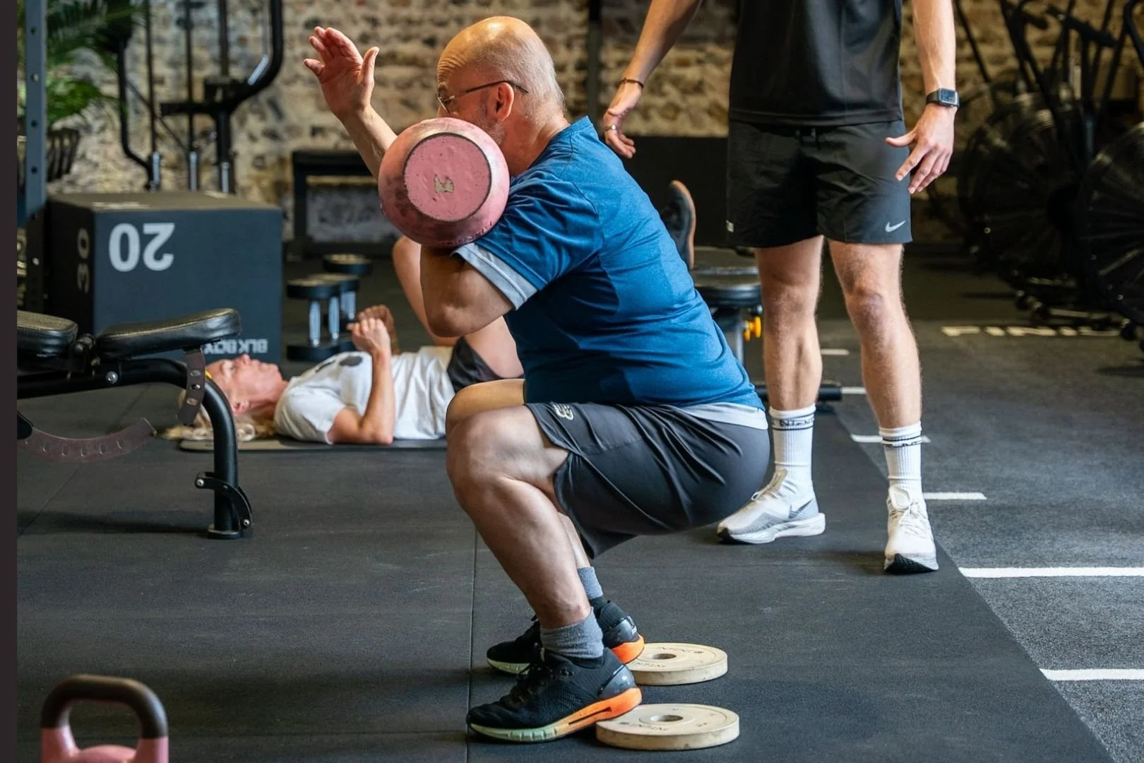 An elderly man with glasses and a blue shirt is squatting while holding a pink kettlebell on his shoulder, in a gym. There is a woman lying on the floor in the background, and another man standing nearby.