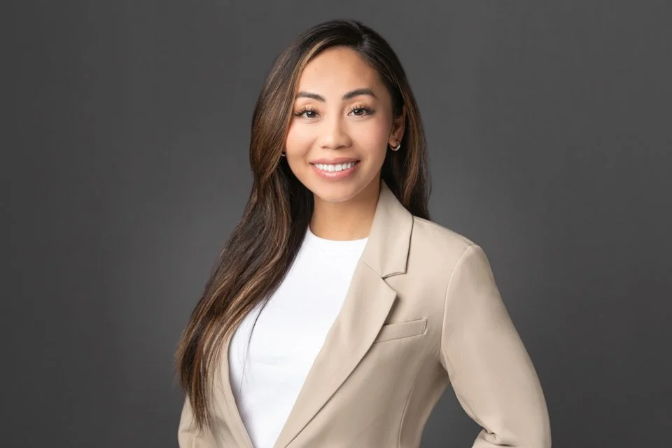 A young woman with long brown hair, smiling, wearing a beige blazer over a white top, standing against a gray background.